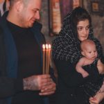 A baby held by mother during an Orthodox candle ceremony indoors with religious icons.